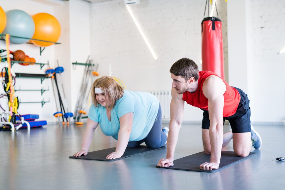 Full length portrait of overweight woman doing fitness exercises in gym with personal trainer helping her, copy space